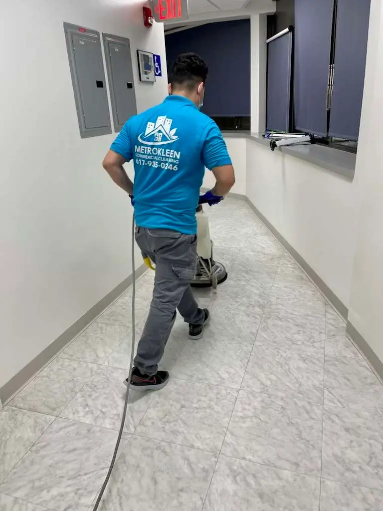 A person in a blue uniform is cleaning a hallway floor with a motorized floor scrubber.